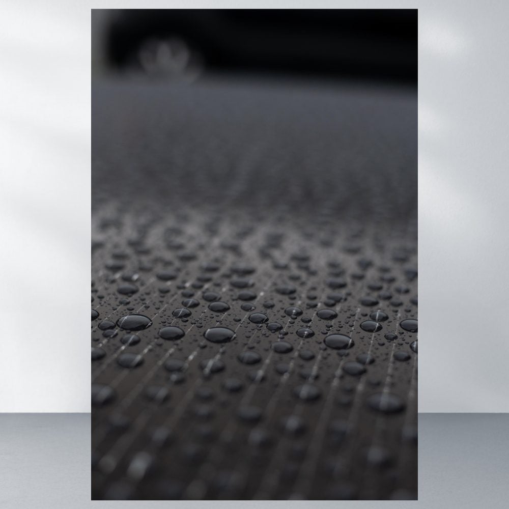 Nahaufnahme von Wassertropfen auf einer dunklen Oberfläche mit einem verschwommenen Auto und einem Trina Vertex S+ 460W Glas-Glas TSM-NEG9R.28 Solarmodul im Hintergrund. Die Tröpfchen bilden Muster, die das Licht reflektieren und die Struktur der Oberfläche betonen.